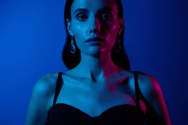 Mavi ve pembe renkli, zarif küpeler takan modern ama dramatik bir havayı çağrıştıran bir kadının yakın çekimi..