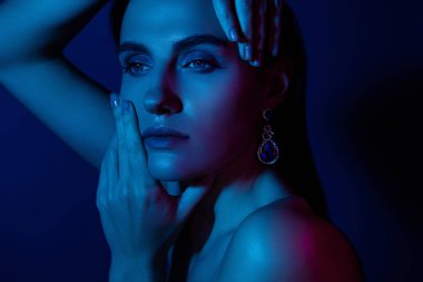 Düşünceli bir ifadeye sahip bir kadın dramatik mavi ışıkla vurgulanır. Zarafetini ve karmaşık küpelerini vurgulayarak gizemli ve büyüleyici bir atmosfer yaratır..