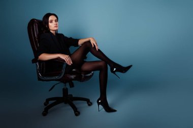 Genç bir kadın, şatafatlı bir siyah ceket ve topuklu ayakkabı sergileyen, çarpıcı mavi bir arkaplana karşı bir sandalyede otururken kendine güven ve zarafet yayıyor..