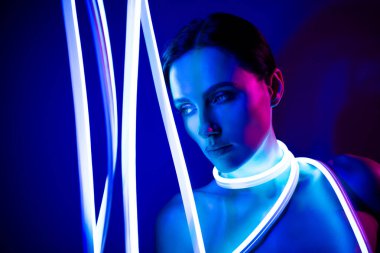 Genç bir kadın karanlık stüdyoda pozlar veriyor, neon ışıkları vücudunu aydınlatıyor, stil ve teknolojinin bir karışımını oluşturuyor..