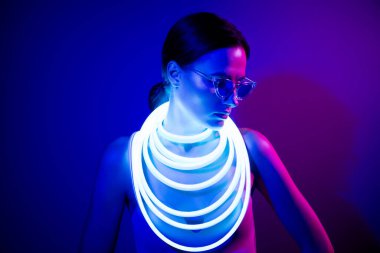 Neon ilmikli kolyesi ve güneş gözlüklü, parlak mavi bir atmosferi olan zarif genç bir kadın. Zarif ve fütüristik bir tarzı var..