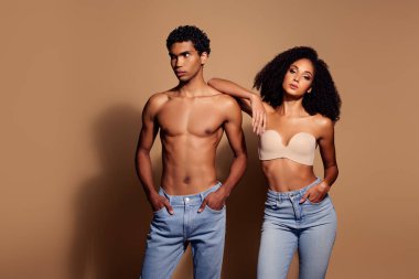 Kot ve iç çamaşırı giymiş zarif genç bir çift stüdyo ortamında özgüven ve tarz yayıyorlar. Bej arka plan onların çarpıcı varlıklarını vurgular.