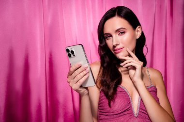 Pembe elbiseli çekici bir esmer, elinde modern bir akıllı telefon tutuyor. Pembe tekstil arka planı lüks ve şık bir görünüm sağlar. Güzellik ve moda temaları için mükemmel bir ortam yaratır..