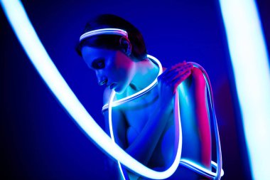 Dramatik bir stüdyo ortamındaki zarif genç bir kadın, neon ışıklı bantlarla gölgesini koyu mavi bir arkaplana vurguluyor..