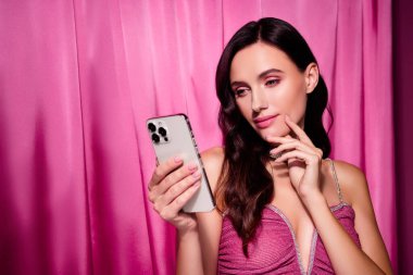 Pembe elbiseli şık bir kadın stüdyo ortamında telefonunu karıştırıyor.