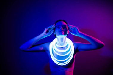 Şık genç bir kadın koyu mavi neon ışıkta güven içinde poz veriyor. Fütürist moda unsurları zarafet ve güzelliği vurguluyor, çekici ve büyüleyici bir görünüm yaratıyor..