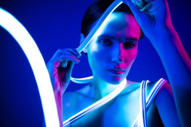 Çarpıcı bir kadın zarif bir şekilde parlak neon ışıklarla poz veriyor, koyu mavi bir zemin üzerine kurulmuş, güzel ve çağdaş moda tarzını sergiliyor..