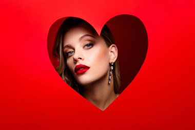 A stylish woman with red lipstick and smoky eye looking through a red heart-shaped cutout, symbolizing passion, love, and sophistication. The vibrant red background enhances the Valentines theme. clipart