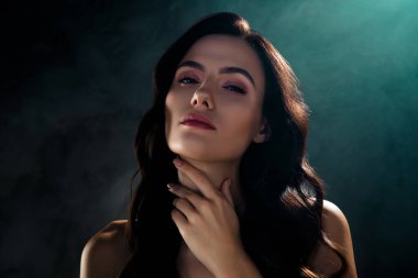 Esmer, alımlı, çekici bir kadının fotoğrafı. Boynuna dokunuyor. Koyu renk arka planda hassasiyet hissediyor..