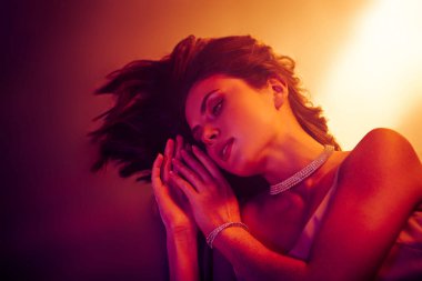 Zarif makyaj ve şık mücevherlerle şık bir kadın, göz alıcı bir atmosfer için canlı, sıcak turuncu ve pembe ışıkla yıkanmış.