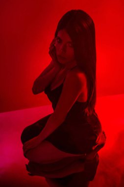 Güzel, genç bir kadın parlak kırmızı ışığın altında poz veriyor. Şık, siyah bir elbise giyiyor. Zarafet ve cazibe saçıyor. Atmosfer stil, moda ve tutkunun büyüleyici temasını vurgular..