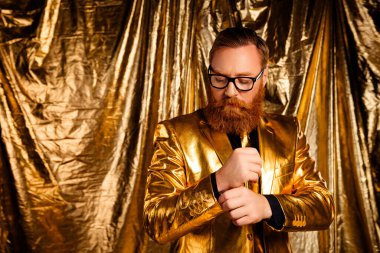 Kırmızı sakallı, parlak altın bir takım giyen, kollarını ayarlayan karizmatik bir adam. Altın arka plan zarif ve göz alıcı havayı arttırır..