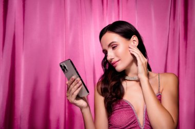Pembe elbiseli şık bir kadın, elinde akıllı bir telefonla canlı pembe bir tekstil geçmişine karşı özgüvenle poz veriyor. Görüntü, bir stüdyo ortamında moda, cazibe ve cazibeyi vurguluyor..