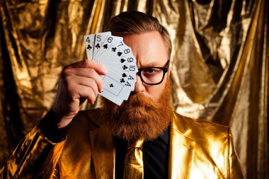 Kırmızı sakallı karizmatik bir adam altın renkli bir arka planda kart oynayarak zarif ve lüks bir atmosfer yaratıyor..