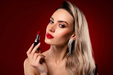 Güzel bir bayan, sarı saçlı ve göz kamaştırıcı bir stili olan parlak kırmızı rujla poz veren canlı bir bordo arka planda.