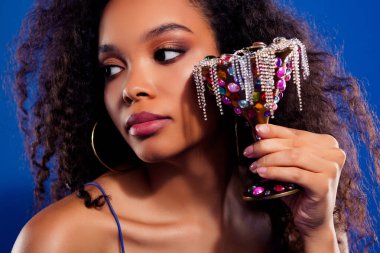 Genç bir kadın mücevherler ve pullarla süslenmiş dekoratif bir kadehle poz veriyor, zarif makyajını ve tarzını vurguluyor, canlı mavi bir zemin üzerine kurulmuş, büyüleyici ve lüks.