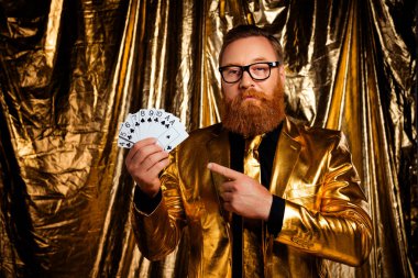 Kızıl saçlı ve sakallı olgun bir adam, parlak altın bir takım elbise giymiş, kendine güvenen bir el kağıt oynuyor, zarafet ve kutlama çağrıştırıyor..