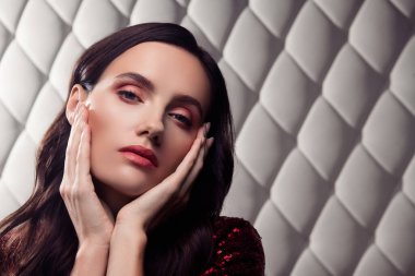 Zarif bir stile sahip genç bir kadın göz kamaştırıcı makyajı ve beyaz örülü bir duvara karşı pullarla süslenmiş zengin kırmızı elbisesi var.