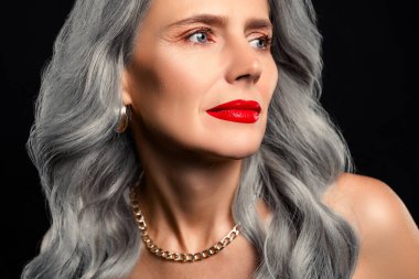 Şık ve olgun bir kadının portresi, göz kamaştırıcı gri saçları, göz kamaştırıcı makyajı ve altın takıları olan zarif ve zamansız bir atmosfer için koyu bir arkaplan üzerine çekilmiş..