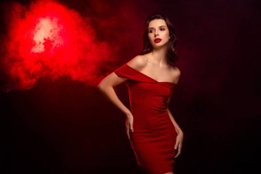 Omuzları açık kırmızı bir elbise giymiş genç bir kadın huysuz, kırmızı ve siyah bir arkaplan karşısında zarif bir şekilde duruyor. Zarafet, tarz ve cazibe sergiliyor..