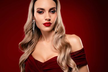Zarif makyajlı, uzun sarı saçlı, koyu kırmızı elbiseli, zarif bir kadın, zengin bir bordo arka planda poz veriyor..