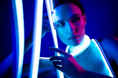 Koyu mavi arka planda parlayan neon ışıklarla poz veren genç kadın, fütüristik stüdyoda zarafet ve modern moda..