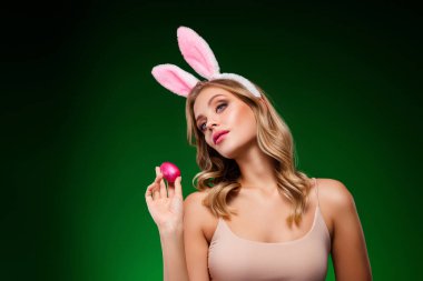 Çekici bir kadın, tavşan kulaklı, zarif bir şekilde dekoratif bir Paskalya yumurtası tutuyor, canlı, yeşil bir tatil arkaplanına karşı kurulmuş, zarafeti, güzelliği, kutlamayı ve şenlik tarzını vurguluyor..