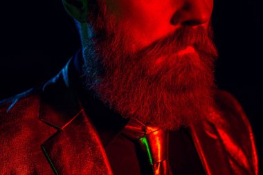 Karizmatik bir erkek figürü, bakımlı sakallı ve parlak takım elbiseli, neon ışıklarla aydınlanan koyu bir arka plan. Gece hayatı ve geçmişe dönük zarafet temaları için mükemmel..