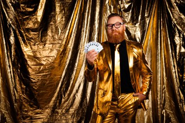 Karizmatik kızıl saçlı, parlak altın takım elbiseli adam elinde kartlar, zarafet ve cazibe ile altın bir zemin tutuyor..