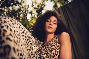 Güzel genç bir kadın, açık havada gün ışığı altında poz veriyor, şık bir leopar deseni giyiyor, yemyeşil arka planda çeşitliliği, zarafeti ve tarzı vurguluyor..