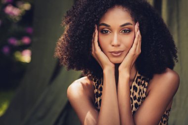 Şık ve kendine güvenen, doğal kıvırcık saçlı bir kadın güneşli bir parkta poz veriyor. Huzurlu bir bahçe ortamında güzellik, çeşitlilik ve zarafet vurgulanıyor..