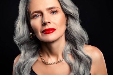 Gri saçlı, koyu kırmızı dudaklı, orta yaşlı sofistike bir kadın. Koyu bir arka fona poz vermiş, zarif ve kendine güvenen bir kadın..