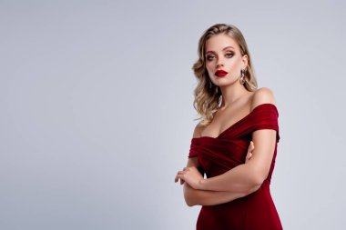 Şık, ciddi görünümlü, kırmızı kokteyl elbiseli bir kadın. Gri renkli arka planda duruyor. Kollarını kavuşturmuş..