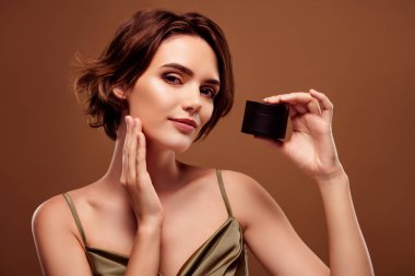 Çekici genç bir kadının fotoğrafı kahverengi arka planda izole edilmiş şık haki rengi kıyafetler giyip krema kavanozu uygula..