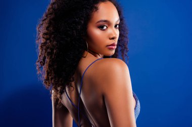 Genç bir kadının zarif portresi omuzlarının üzerinden bakıyor, parlak mavi bir arkaplan üzerine kurulu, kıvırcık saçlarını ve büyüleyici tarzını sergiliyor..