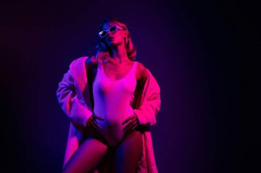 Güzel, genç bir kadın, neon pembe ve koyu mor bir ışıkla kendine güvenerek poz verir. Cazibesini ve zarafetini bir beden ve şık bir ceketle sergiler..