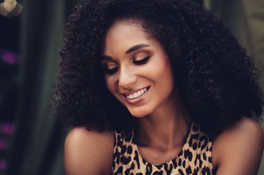 Genç, doğal olarak güzel bir kadın. Parlak, kıvırcık saçlı ve şık giyinmiş. Açık hava ortamında gün ışığında..