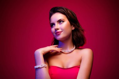 Zarif bir esmer kadın makyaj ve mücevher giyiyor, büyüleyici bir ifadeyle zarif poz veriyor, canlı pembe ve kırmızı gradyan arka planda..