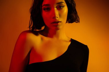 Büyüleyici özellikleri olan genç esmer bir kadın gradyan turuncu renkli arka plana karşı poz veriyor, çağdaş moda tarzı ve şehvetini sergiliyor..