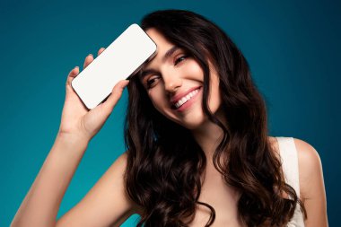 Neşeli bir kadının elinde parlak mavi bir arka plan karşısında boş ekranlı bir akıllı telefon var. Teknoloji, güzellik veya reklam teması için ideal.
