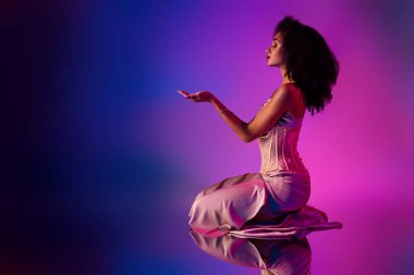 Moda ve güzel bir genç kadının stilize edilmiş stüdyo fotoğrafı. Zarif bir kıyafet giyerek, neon ışıklı bir ortamda zarif bir şekilde diz çöker, kendine güveni ve duruşunu gösterir..