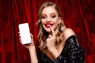 Güzel bir kadın telefonunu sergiliyor, zarif ve alımlı kıyafetleriyle lüks kırmızı kadife arka planda, cazibe ve entelektüelliği vurguluyor..