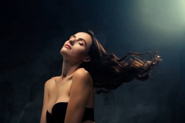 Koyu renk saçları olan zarif bir genç kadın, koyu bir arka plana karşı zarifçe poz verir, çarpıcı ışıklandırmayla aydınlatılır, sofistike ve güzel bir hissi uyandırır..