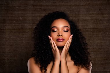 Zarif, kıvırcık saçlı, güzel makyajlı ve pürüzsüz ciltli genç bir kadın dokulu kahverengi bir arka plan önünde özgüvenle poz veriyor, cazibe ve incelik sergiliyor.