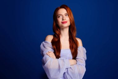 Genç, çekici, kızıl saçlı, açık mavi yumuşak bir elbise giyen, kollarını minimalist mavi bir arka plana çarpıştırarak, şık ve zarif bir görünüm sergileyen bir kadın..