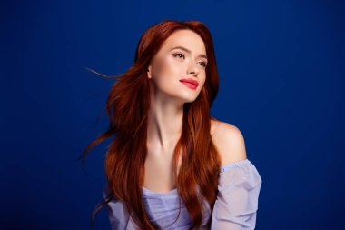 Uzun kızıl saçlı çarpıcı bir genç kadın zarif poz veriyor, canlı mavi arka planlı bir moda fotoğrafında güzelliğini ve tarzını sergiliyor..