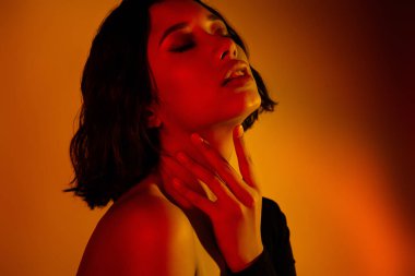 Kısa siyah saçlı genç bir kadın canlı turuncu bir zemin altında dramatik bir ışıkla zarifçe poz veriyor. Dekor onun duygusal ifadesini vurguluyor, resme modern ve sanatsal bir yaklaşım..