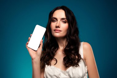 Zarif esmer kadın kameraya bakıyor, elinde boş bir akıllı telefon ekranı, modern mavi bir gradyan zemin üzerinde güzelce tasarlanmış, teknoloji, moda ve sofistike temalar çağrıştırıyor..