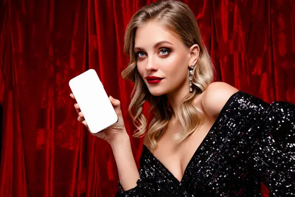 Göz kamaştırıcı makyajı ve şık elbisesi olan güzel sarışın bir manken elinde telefon, lüks bir kırmızı kadife arka planda sergileniyor, büyüleyici ve sofistike..