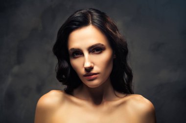 Koyu bir arka planda zarif bir ifadeye sahip genç esmer bir kadının yakın çekimi. Görüntü, onun parıldayan ten rengini ve varlığını, kendine olan güvenini ve doğal güzelliğini vurgular..